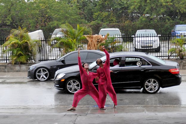 Two side-by-side performers in hot pink lace jumpsuits bend their knees and look up towards their raised arm in a rainy street in front of a parking lot with palm trees and green foliage. Behind them a person wearing a pink headmask drives a car with a speaker in the back window. 