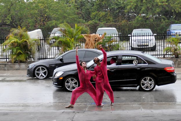 Two side-by-side performers in hot pink lace jumpsuits bend their knees and look up towards their raised arm in a rainy street in front of a parking lot with palm trees and green foliage. Behind them a person wearing a pink headmask drives a car with a speaker in the back window.