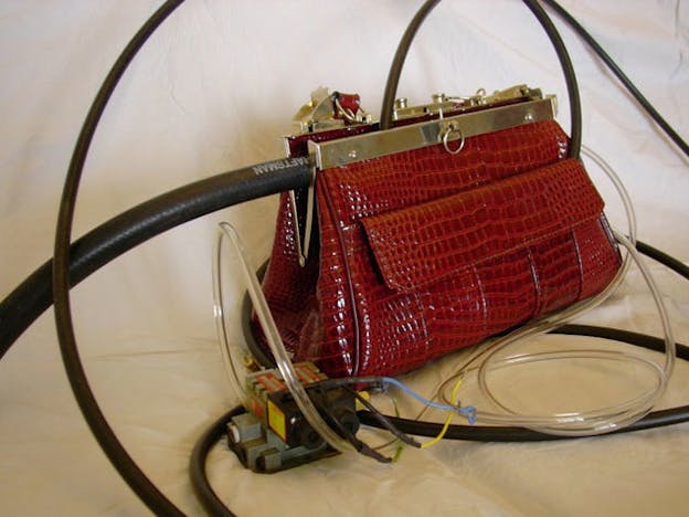 A close up image of a red alligator skin purse with wires coming out of it in front of a white cloth background. 