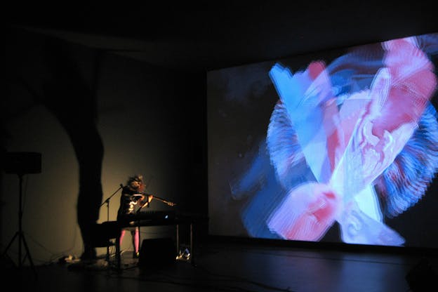 A dim lit space has a projection on the entire wall of pink and blue swirls. On the other wall a figure surrounded by multiple musical instruments plays a string instrument.