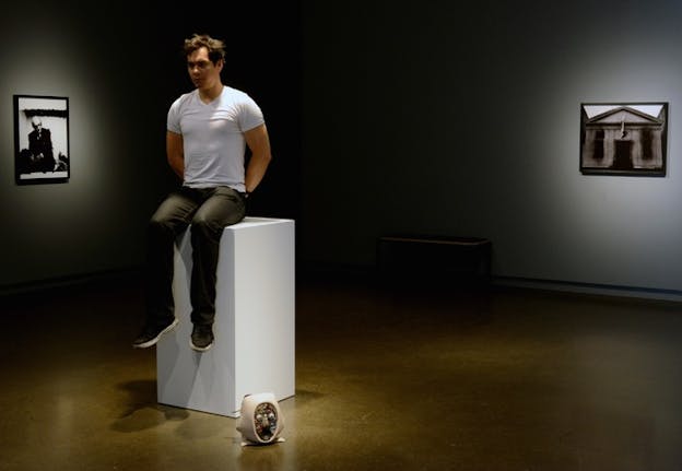 A person in a white shirt and dark jeans sits on top of a an art stand, with their arms on their back. At the end of their feet an open face sculpture.  