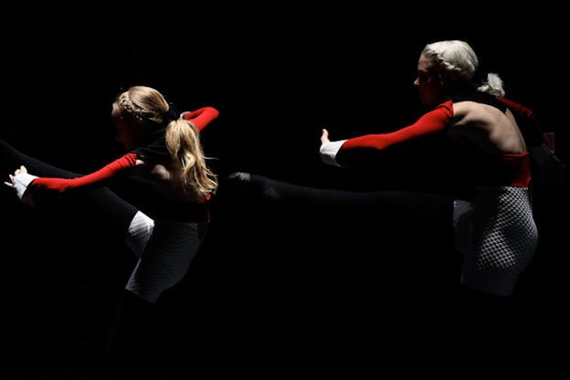 Two performers stand in a line within a darkened room. They are hunched over with their right leg lifted directly in front of them and their left arm lifted parallel to their leg. They wear their hair in braids and have costumes of red leotards, white shorts, and black tights. 