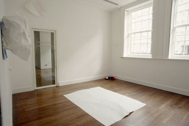 A white sheet lays on top of something on the wooden floor of an otherwise bare white room. Two windows on the right wall illuminate the space with natural light. A white object hangs on the left wall.