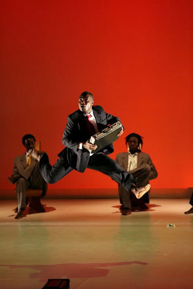 One performer wearing a black tuxedo outfit leaps in the air carrying a briefcase. In the background, two other performers both wearing khaki colored tuxedo outfits are in a half-kneeling position. The stage has a baby yellow colored floor and a red backdrop.