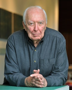 Portrait of Jasper Johns leaning forward with intertwined fingers on the edge of a surface. He wears a blue button-up - gray hair, strong brows, and blue eyes.