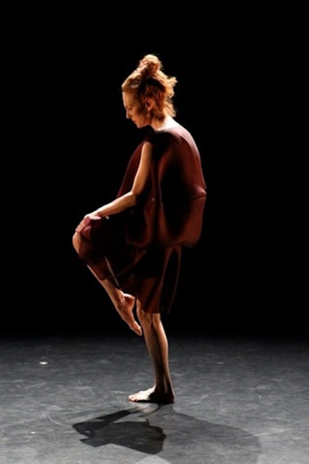 Performer wearing a flowy wine colored dress creates a triangle-shape by raising one pointed toe to their bent knee and looking down at the floor of a gray stage with a pitch black backdrop.