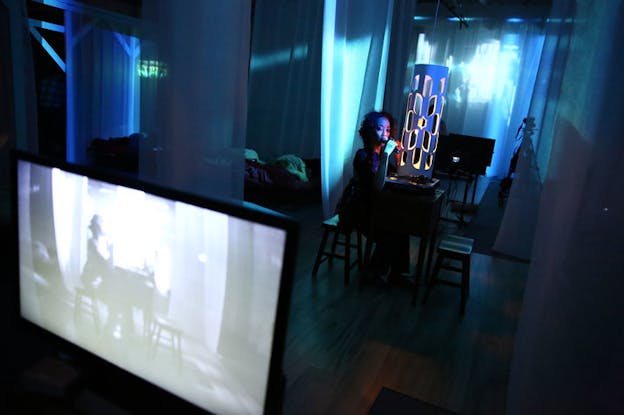 Performer holds a microphone to their mouth sitting at a table in the center of a translucent draped glowing blue-lit room. At the bottom left of the frame a television screens portrays the performer.