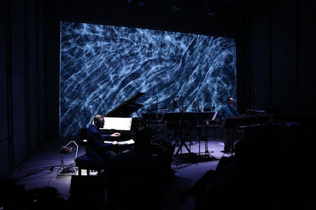 Winat and a pianist perform in a dark room in front of a swirling white and ocean-blue screen projection.