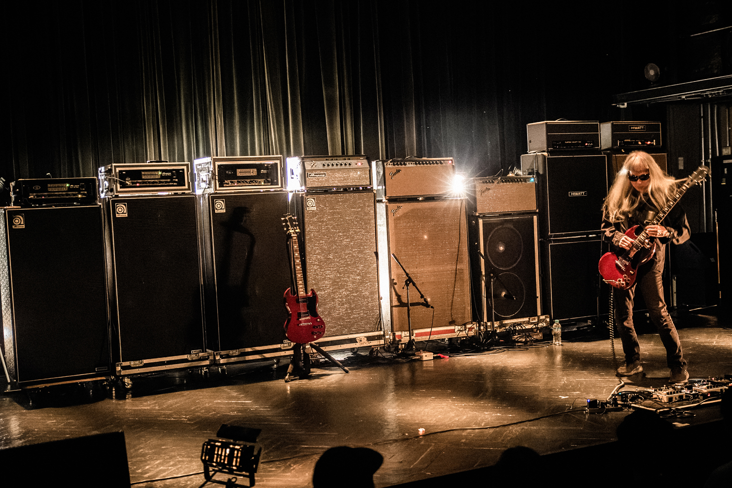 Keiji Haino stands on a black stage in front of a row of amplifiers playing a cherry red Gibson SG guitar on the far right side of the image, his hips thrust slightly forward.