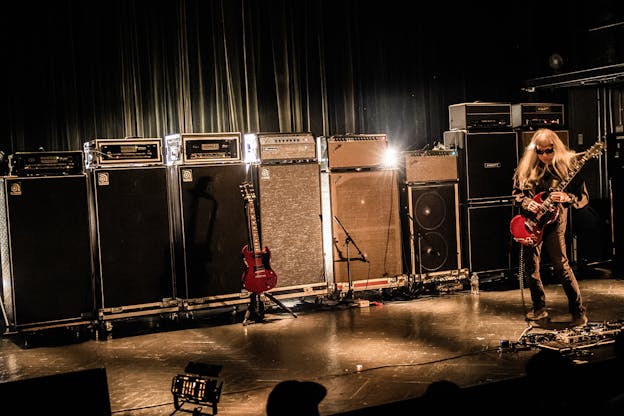 Keiji Haino stands on a black stage in front of a row of amplifiers playing a cherry red Gibson SG guitar on the far right side of the image, his hips thrust slightly forward.