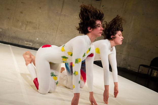 Two performers dressed in white leotards with colors and fruit patterns adorning each, stand on all fours with their hands supported by their fingers, as they throw their heads back resulting in their short brown hair to fly upwards. 