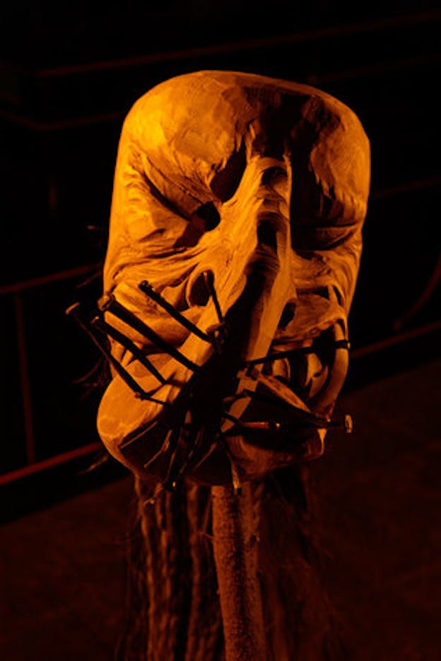 A close up of one wooden sculpture carved with a face with squinting eyes, a long nose, and a mouth which is grimacing, bearing sharp teeth. On the bottom of the nose, numerous large nails are hammered into the sculpture at different angles. The sculpture is held up by a wooden branch and in the background, blurred, long hair is visible. 