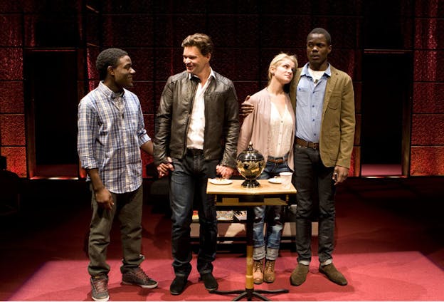 Four persons stand behind an urn on a small table in a room with a red floor and red walls, linking hands and making varying expressions at each other.