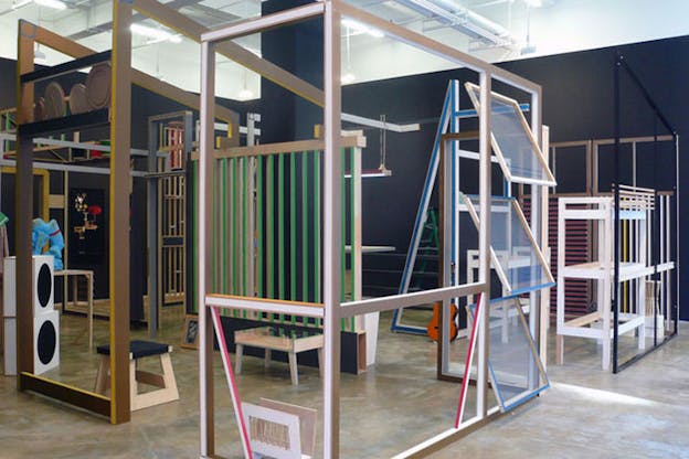 An installation image of several multi-colored wooden frame structures. In the center, there is a black square column and the walls are black. Some assorted items adorn the structure including a small orange guitar and some brown plates. 