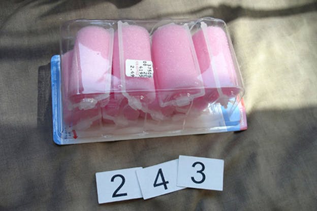 Pink foam curlers are in a plastic container on a grey surface. Three small papers each with the number 2, 4, and 3 printed on them in a black font lay in front of the curlers.