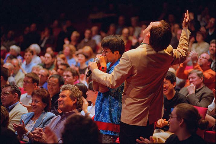Two people stand up out of the audience chairs. One of them wears a blue shirt and speaks closely into a microphone. The other wears a tan suit jacket and leans back with their arms up.