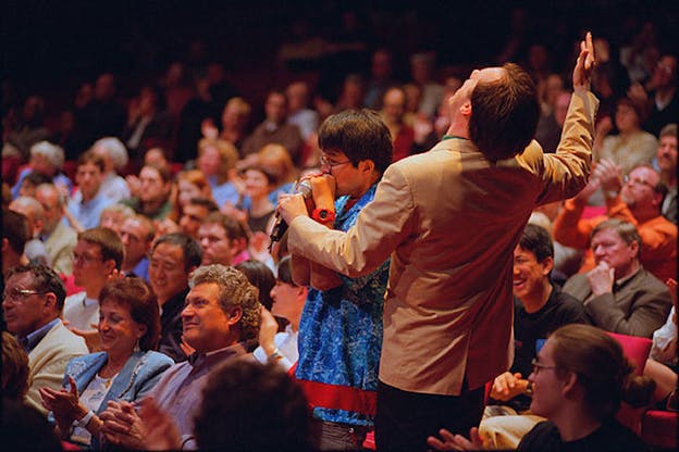 Two people stand up out of the audience chairs. One of them wears a blue shirt and speaks closely into a microphone. The other wears a tan suit jacket and leans back with their arms up.
