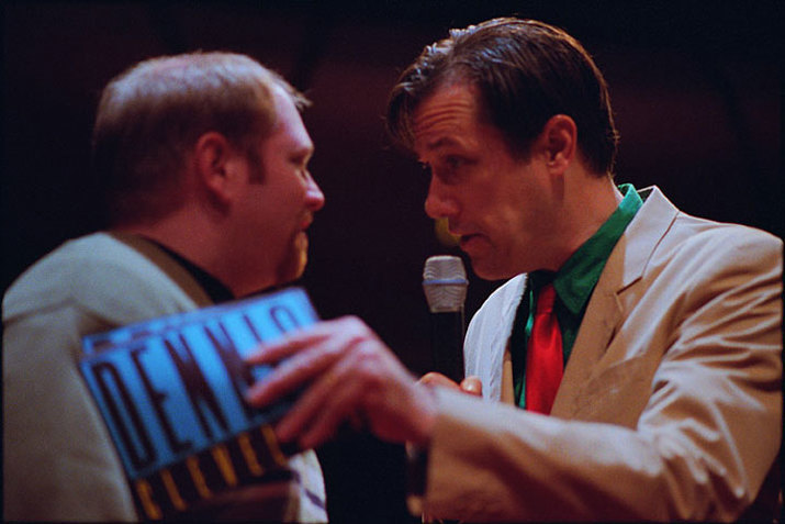 A close up shows two performers facing each other. The one on the right wears a beige suit jacket, green shirt, and bright red tie. They hold flyers saying Dennis Cleveland which they press against the other performer's shoulder. Looking intently at that performer, they speak into a microphone. The performer on the left wears a ligh green top and looks back at the other performer.