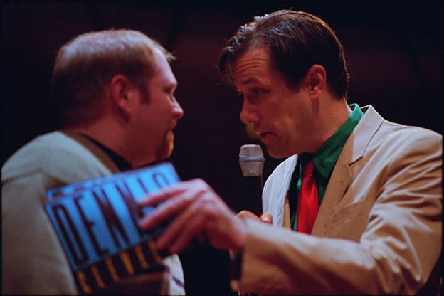 A close up shows two performers facing each other. The one on the right wears a beige suit jacket, green shirt, and bright red tie. They hold flyers saying Dennis Cleveland which they press against the other performer's shoulder. Looking intently at that performer, they speak into a microphone. The performer on the left wears a ligh green top and looks back at the other performer.