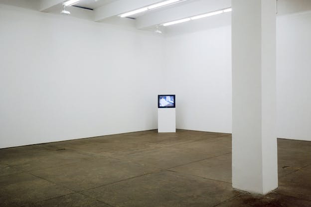An empty gallery room with white walls and beige floor. In the far back corner an open TV situated on a white small column.