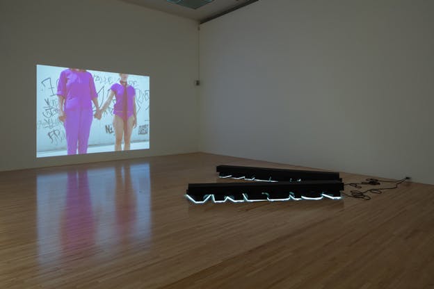 A projection on a wall of two figures dressed in purple holding each other hands, their heads not visible. Two long black structures sit on the floor, lights coming from underneath them and cables on connected to sockets on the wall. 
