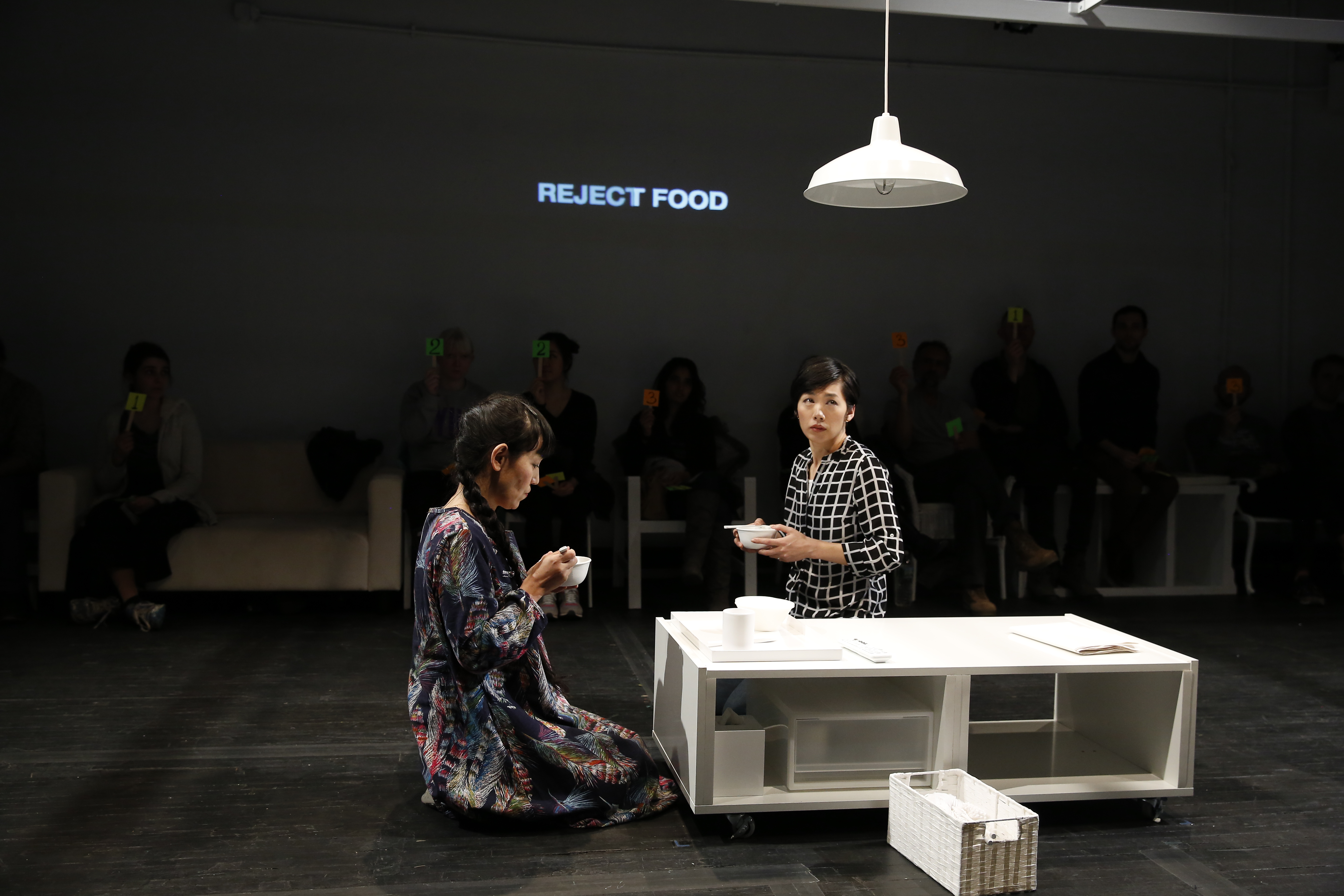 Two performers kneel on the ground in front of a low-lying white rectangular table. They each hold a white bowl. The performer on the left wears a dress decorated with feathers and looks down at their bowl. The second performer’s body is facing the first, but their eyes are looking up and to the side. The words “REJECT FOOD” are projected on the wall behind them. Audience members sit along the outer edges of the room holding numbers written on brightly colored paper and affixed to wooden handles.
