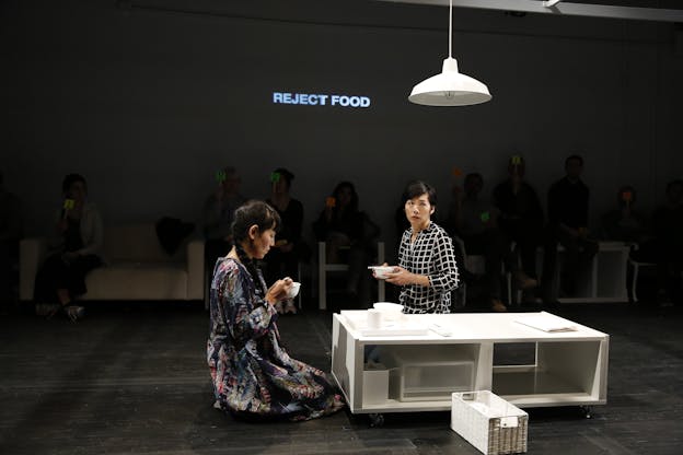 Two performers kneel on the ground in front of a low-lying white rectangular table. They each hold a white bowl. The performer on the left wears a dress decorated with feathers and looks down at their bowl. The second performer’s body is facing the first, but their eyes are looking up and to the side. The words “REJECT FOOD” are projected on the wall behind them. Audience members sit along the outer edges of the room holding numbers written on brightly colored paper and affixed to wooden handles.