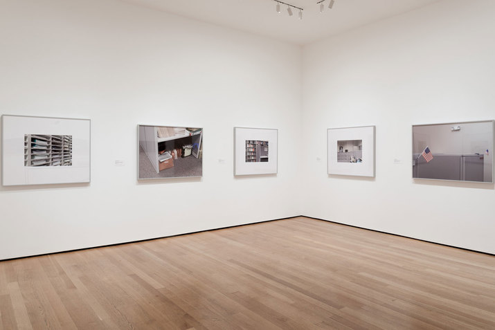 Blank white walls display framed gray-toned photographs of office shelves, a cubicle, a filing cabinet, a locker, and a miniature American flag.