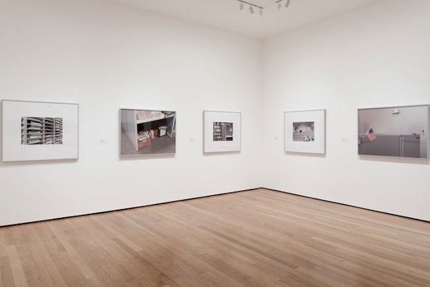 Blank white walls display framed gray-toned photographs of office shelves, a cubicle, a filing cabinet, a locker, and a miniature American flag.