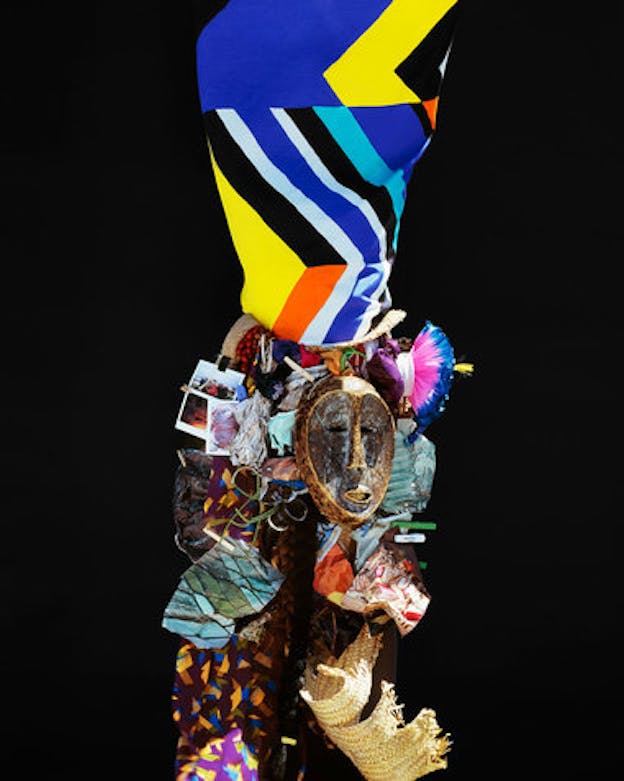 Collage sculpture of mixed materials including bright-blue, orange, and yellow chevron-striped fabric, a wooden face mask, torn woven straw, and polaroids against a pitch black backdrop.