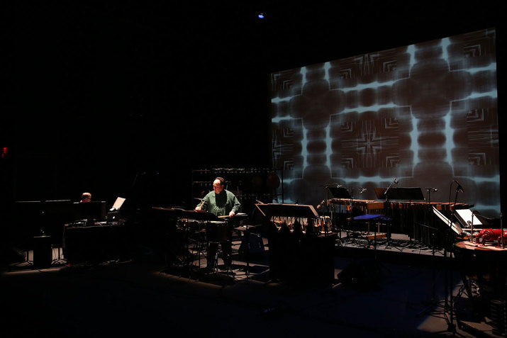 Winant and a fellow performer on a dimly lit stage, surrounded by an array of percussive instruments. On the back wall, a screen projects a blue and black kaleidoscopic pattern. 