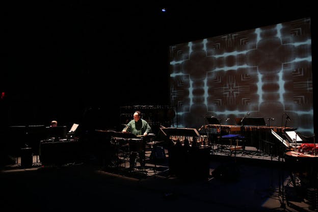 Winant and a fellow performer on a dimly lit stage, surrounded by an array of percussive instruments. On the back wall, a screen projects a blue and black kaleidoscopic pattern. 
