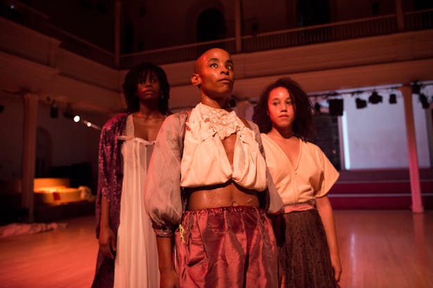 Three performers stand in formation in the center of the image facing slightly to the right and wearing loosely fitted garments. They are bathed in a soft red/pink light. Jasmine and Angie look upward; Dominica gazes down and to the left.