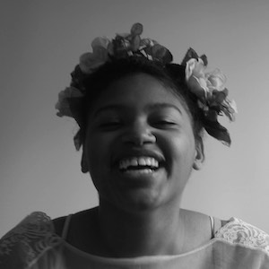 Black and white portrait of Alijah Webb in the middle of laughter, wearing a flower crown and a flower-detail white top.