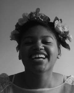 Black and white portrait of Alijah Webb in the middle of laughter, wearing a flower crown and a flower-detail white top.