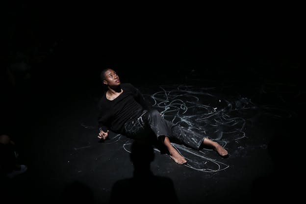 A performer wearing all black leans on a black floor and looks up, scared. In the middle of the floor, looping lines have been drawn in white chalk which overlap onto the performer's pants. The shadow of the torso of a person oriented perpendicular to the performer is visible in lower middle.  