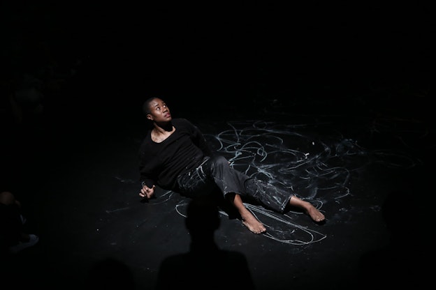 A performer wearing all black leans on a black floor and looks up, scared. In the middle of the floor, looping lines have been drawn in white chalk which overlap onto the performer's pants. The shadow of the torso of a person oriented perpendicular to the performer is visible in lower middle.  