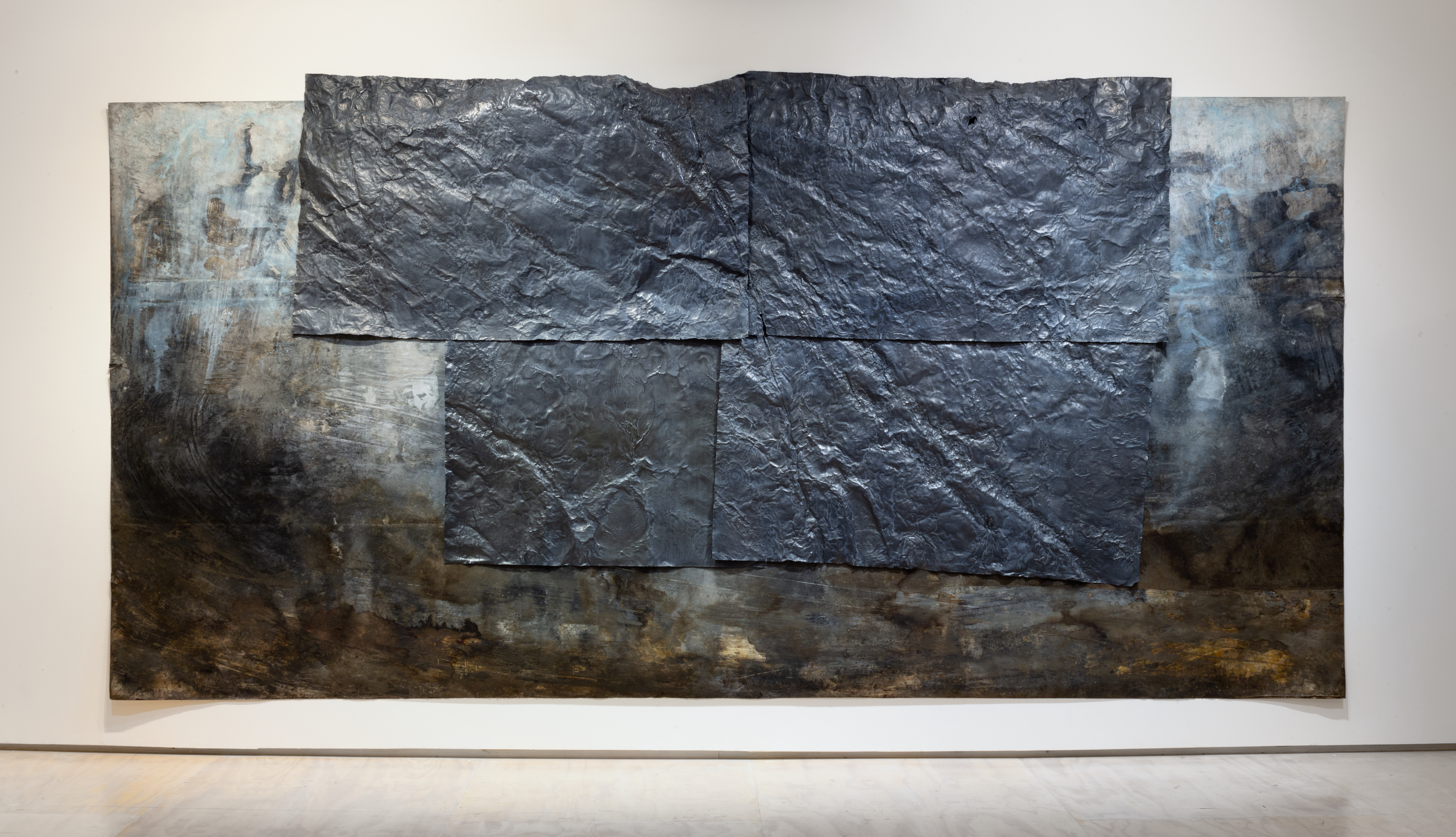 A long rectangular painting with dark hues of black, brown, and tan concentrated toward the horizontal edges and bottom, with white and blue toned areas towards the top. Centered and aligned with the top edge of the painting four dark grey, crinkled, metallic sheets are fixed to the front of the painting.