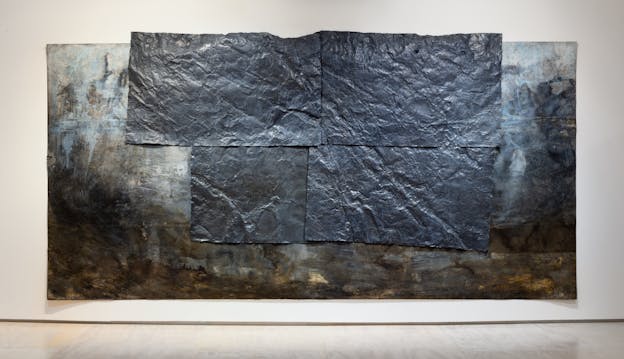 A long rectangular painting with dark hues of black, brown, and tan concentrated toward the horizontal edges and bottom, with white and blue toned areas towards the top. Centered and aligned with the top edge of the painting four dark grey, crinkled, metallic sheets are fixed to the front of the painting.