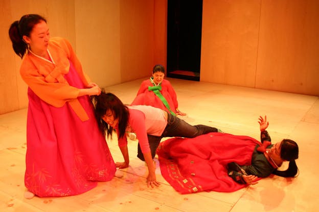 Three people dressed in traditional Korean wear are in a fight with one person dressed in modern wear. One of them grabs the girl's hair while the other two fall to the ground. The set has wood floors and walls and an open doorway to darkness in the back right corner.