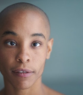 Jasmine Hearn gazes into the camera, their head shaved and lips slightly parted. They are in sharp focus against a grey blue background.