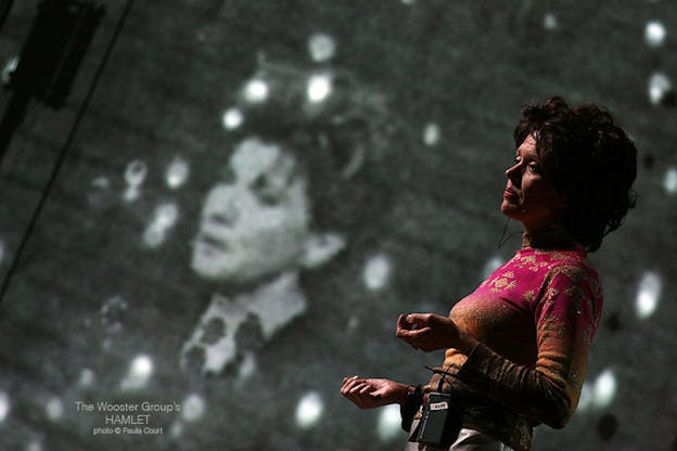 Kate Valk is in front of a projection of a person in black and white. Kate Valk looks somber and wears a pink and gold patterned long sleeve top. She holds her hands limply at her waist.