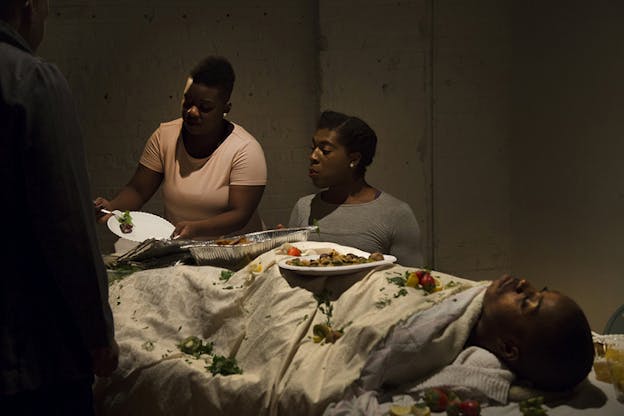 A figure covered by a white sheet lies down on a surface while other people discard food from paper plates and aluminum trays on her.