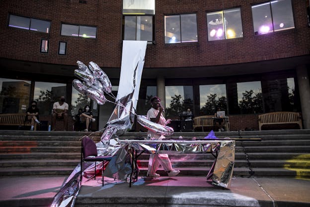 Autumn Knight performs with a table covered ina metallic sheet, metallic balloons attached to her body, and a long metallic sheet hung from the buliding behind her.  A bright pink light reflects on the performer and the balloons. She holds a microphone and walks in mid-stride across the courtyard. Audience sit up the stairs behind Autumn and watch.