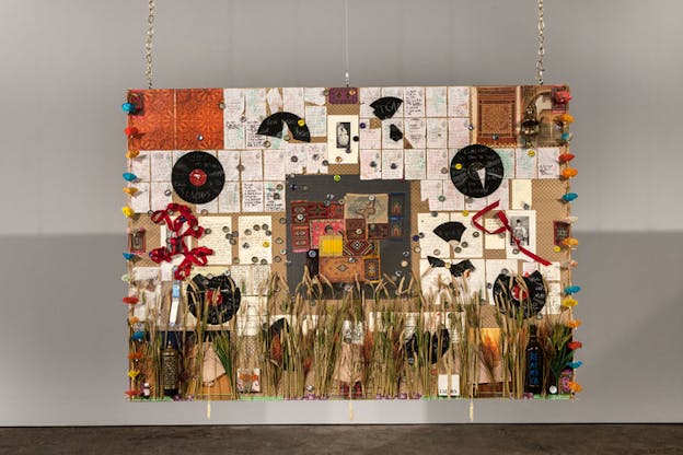 Hanging quilt with arranged mixed material: pages of handwritten notes, broken record pieces, fragments of costumes and props, fabric strips, paper parasols, and along the bottom edge beside a bottle painted with the word 
