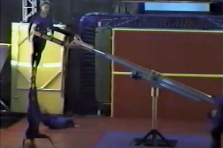 A gymnast supported by mechanism surrounding his waist balances his upside down form by standing with his hands on the feet of another gymnast who lies his upper body down.