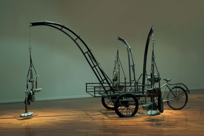 A side view of a mixed media installation made of found and recycled objects. A tricycle is used as the base to support one longer and two shorter curved iron structures branching outward, each with several river stones are hung at the end. The installation sits on a wooden floor against a palladian blue background with natural lighting coming from above.