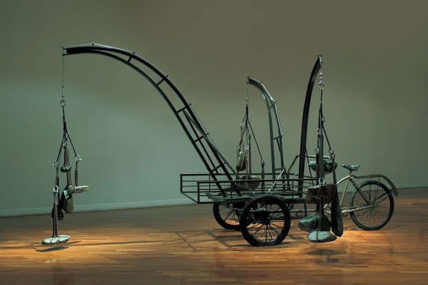 A side view of a mixed media installation made of found and recycled objects. A tricycle is used as the base to support one longer and two shorter curved iron structures branching outward, each with several river stones are hung at the end. The installation sits on a wooden floor against a palladian blue background with natural lighting coming from above.