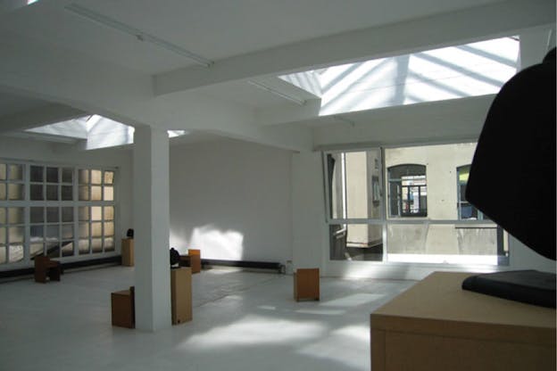 An interior image of a bare white room filled with only a few brown boxes. There are large windows on the far wall which look out onto windows in a building next door. Large skylights illuminate the room. 