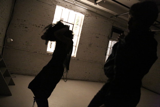 Two shadowed figures in movement stand inside a white room, sideways to the viewer.